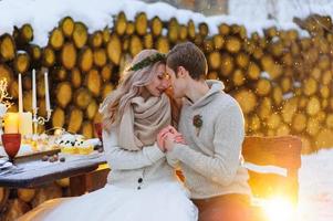 Bräutigam küsst seine Braut an der Schläfe. Jungvermählten mit Blumenstrauß sitzt auf Schnee auf dem Holzhintergrund. Winterhochzeit foto