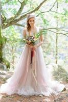 glückliche Braut in einem rosa Hochzeitskleid. das mädchen hält einen hochzeitsstrauß in ihren händen. Hochzeitszeremonie im Boho-Stil im Wald. foto