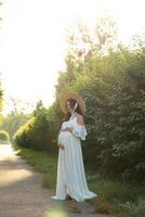 Schwangere Frau mit Hut, die in einem Kleid auf einem Hintergrund von grünen Bäumen posiert. foto