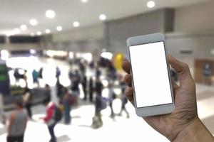 Hand, die ein Smartphone mit weißem, leerem Bildschirm und verschwommenem Wartebereich für Gepäck im Hintergrund des Passagierterminals hält foto