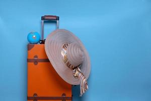 Orangefarbener Koffer mit Mini-Globus und Sommerhut isoliert auf blauem Hintergrund für Reisekonzept mit minimalem Stil foto