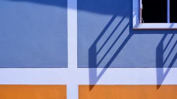 sonnenlicht und schatten auf der oberfläche des weißen holzfensters mit blau und orange bemaltem wanddekorationshintergrund, außenarchitekturkonzept foto