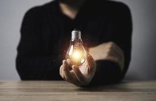 Geschäftsmann, der eine leuchtende Glühbirne mit orangefarbenem Lichtstrahl auf einem Holztisch hält, um kreative Denkideen und zukünftiges Innovationstechnologiekonzept zu inspirieren.