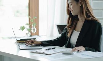 Schöne asiatische Frau, die im Büro mit Laptop und Taschenrechner sitzt. glückliche Geschäftsfrau, die an einem Schreibtisch in einem Büro mit einem Tablet-Computer sitzt. foto