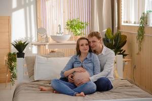 Schwangeres Paar umarmt sich, während es auf dem Bett sitzt foto