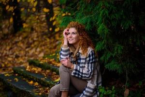 schönes rothaariges Mädchen mit lockigem Haar und blauen Augen. Das Mädchen trägt eine karierte Jacke. Das Mädchen sitzt auf einer Bank. foto