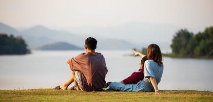 mutter und zwei söhne stehen neben einem großen see und sehen im hintergrund bergblick, mutter zeigt mit dem finger auf den wald. Idee für gemeinsame touristische Familienreisen bis hin zum Outdoor-Trip foto