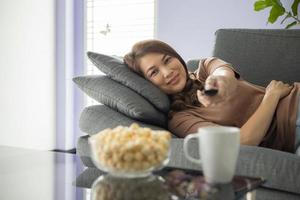 asiatische frau, die fernbedienung hält und mit glück auf den fernseher schaut, während sie auf einem gemütlichen sofa mit popcorn und einem glas getränk zu hause im wohnzimmer liegt foto