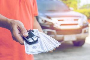 Mann hält Autoschlüssel und Dollar mit Auto im Hintergrund foto