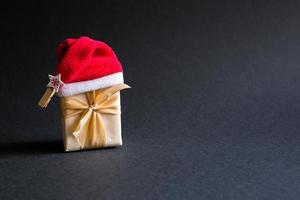 eine schachtel mit einem weihnachtsgeschenk in einem paket, das mit einem band und einer weihnachtsmütze auf schwarzem hintergrund gebunden ist. schwarzer freitag, geschenke für das neue jahr kaufen. Platz für Text foto