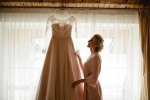 Braut im Nachthemd, das ihr Hochzeitskleid im Schatten hält foto