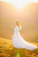 Frau im Hochzeitskleid läuft über das Feld in Richtung der Berge foto
