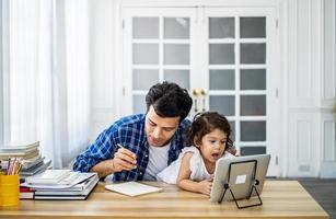 Junger attraktiver Vater und kleine süße Tochter, die sich das Video-Tutorial auf dem Tablet ansehen und zu Hause mit Freude Hausaufgaben unterrichten. foto