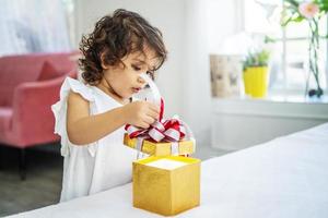 Porträt eines entzückenden kleinen Mädchens, das ein Geburtstagsgeschenk öffnet und mit einem überraschten, freudigen Gesichtsausdruck nach innen schaut