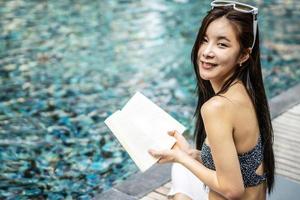 schöne junge asiatische frau sitzt am rand des pools im freien. Sie liest ein Buch, während sie sich in der Nähe des Swimmingpools entspannt