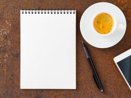 sauberes weißes blatt in einem offenen spiralgebundenen block, stift, handy und tasse kaffee auf dem eisen des rostigen metalltisches, draufsicht foto