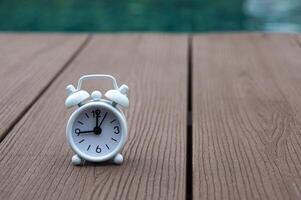 Weißer Wecker auf Holzboden mit verschwommenem Swimmingpool-Hintergrund. die Uhr auf 9 Uhr gestellt. foto