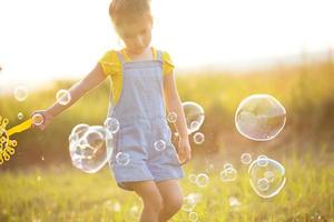 ein mädchen in einem jeansoverall bläst im sommer auf einem feld bei sonnenuntergang seifenblasen. internationaler kindertag, glückliches kind, outdoor-aktivitäten. sommer hintergrund. gesunder und umweltfreundlicher Lebensstil foto