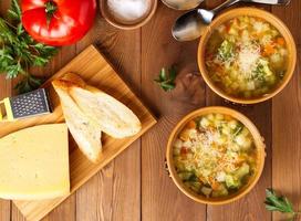 zwei Schüsseln Minestrone-Suppe mit Käse auf Schneidebrett und Gemüse foto