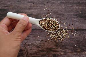 Nahaufnahme der Hand, die den weißen Suppenlöffel mit Quinoa-Samen auf Holztischhintergrund hält. Quinoa ist eine gute Proteinquelle für Menschen, die sich pflanzlich ernähren. Ansicht von oben. foto