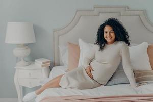 lächelnde junge schwangere Mischlingsdame, die auf dem Bett ruht und die letzten Monate der Schwangerschaft genießt foto