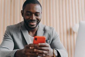 Fröhlicher junger afroamerikanischer Mann, der online auf dem Smartphone chattet und mit einem Lächeln auf den Telefonbildschirm schaut foto