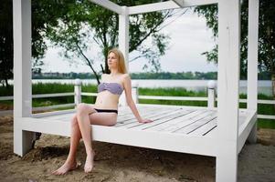 Porträt einer fabelhaften jungen Frau im Bikini, die auf einem weißen Holzpavillon im Park neben dem See sitzt und posiert. foto