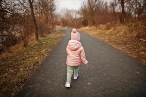 Rückseite des laufenden Babymädchens in rosa Jacke auf dem Weg. foto