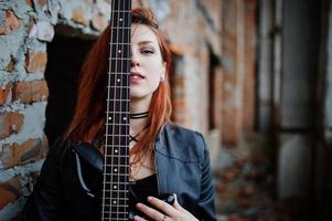 Rothaarige Punkmädchen tragen auf Schwarz mit Bassgitarre an einem verlassenen Ort. Porträt einer gotischen Musikerin. nahaufnahme gesicht der schwärze person mit gitarrenriff. foto