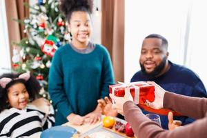 afroamerikanische familie überrascht zusammen mit einem geschenk am weihnachtstag beim abendessen. foto