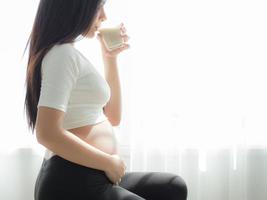 eine schöne schwangere Frau, die steht, ihren Bauch hält und Sojamilch trinkt