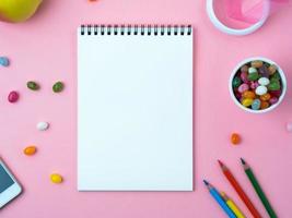 offenes notizbuch mit einem sauberen weißen blatt, süßigkeiten, handy, kreide, dekorationen auf einem rosafarbenen hellen tisch