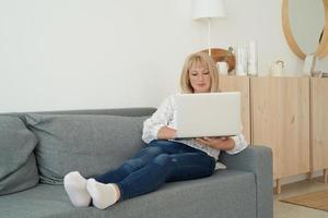 Reife Frau mit Laptop, während sie auf einem bequemen Sofa sitzt, Wohngebäude foto