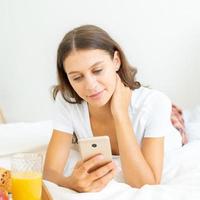 ziemlich weiblich aussehende soziale Medien auf dem Handy, das im Bett liegt. natürliche realistische Schönheit foto