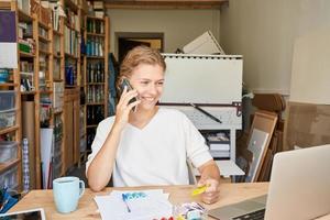 junger weiblicher Unternehmerdesigner, der am Handy spricht und auf Laptop schaut und lächelt foto