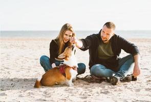 junges glückliches paar von mann und frau mit corgi-hund, der am sand sitzt. zwei personen, männliches fütterndes haustier foto