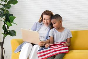 großmutter und enkel spielen fröhlich im wohnzimmer, frau schaut auf laptop-bildschirm und junge hält usa-flagge