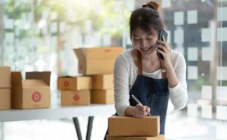 glückliche junge asiatische geschäftsinhaberin bereitet paketbox vor und überprüft online-bestellungen von produkten für die lieferung an den kunden auf dem smartphone. Online-Shopping-Konzept. foto