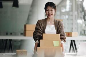 glückliche junge asiatische frau start kleinunternehmen freiberuflich tätig hält paketbox und computer laptop. lieferkonzept für online-marketing-verpackungsboxen foto