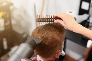 Der Friseur trocknet und stylt die Haare mit Hilfe eines Haartrockners und eines Kamms für einen Schuljungen, der den Friseur während einer Coronavirus-Pandemie besucht. foto