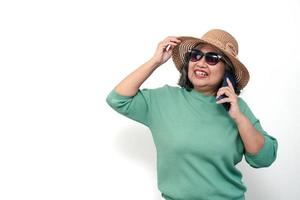 Porträt einer glücklichen asiatischen Seniorin, die eine Sonnenbrille, einen Hut trägt und ein Smartphone hält, das isoliert auf weißem Hintergrund steht. konzept des touristischen genießens und des spaßes am valentinstag nach der pensionierung foto