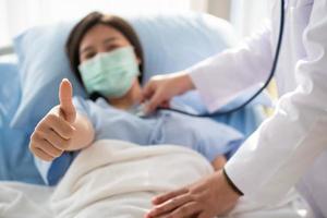 glückliche asiatische patientin trägt eine maske, liegt auf dem bett und hebt den daumen hoch. wenn ein Arzt mit einem Stethoskop die Lunge abhört. konzept des glaubens an behandlung und versicherungsschutz
