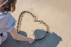 Handfrau, die Herzen auf dem Sandstrand an einem schönen Meerblickhintergrund zeichnet. foto