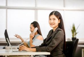 lächelnde asiatische offizierfrau entspannt sich im büro und schaut in die kamera, junge geschäftsfrau sitzt am schreibtisch, geschäftsleute im büro foto