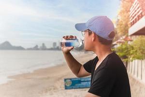 Mann, der Wasser aus einer Flasche trinkt, sitzt draußen neben dem Strand. foto