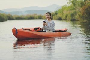 jüngere asiatische frau mit smartphone in der hand, die im seekajak sitzt und vor glück lächelt foto