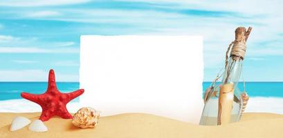 Weißes Papier, das im Sand des Strandes steckt, umgeben von Seesternen, Muscheln und einer Flasche mit einer Botschaft. saubere oberfläche für werbetext foto