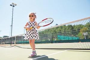 süßes kleines Mädchen, das draußen auf dem Tennisplatz Tennis spielt foto