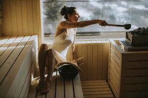 junge Frau gießt Wasser auf heißen Stein in der Sauna foto