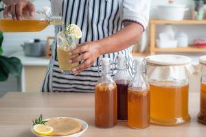 die hand des kochs hält eine flasche und ein glas hausgemachten fermentierten kombucha-tee, abwechslungsreiche aromen in flaschen und gläsern, die mit obst, scoby auf holztisch gemischt werden. Gesundes natürliches probiotisches Getränk. foto